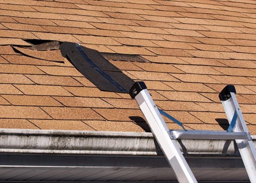 A ladder rests on the gutters of a home with a damaged shingle roof.