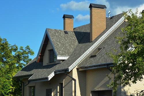 A house with a pitched roofing construction with asphalt shingles installed, attic windows, skylights, soffit and fascia boards, ventilation, two brick chimneys, and roof gutters.